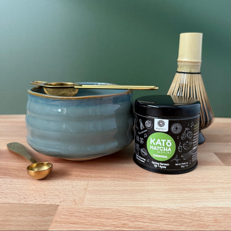 Tea-making set with a blue ceramic bowl, gold whisk, scoop, and Kato Matcha container on a wooden surface.