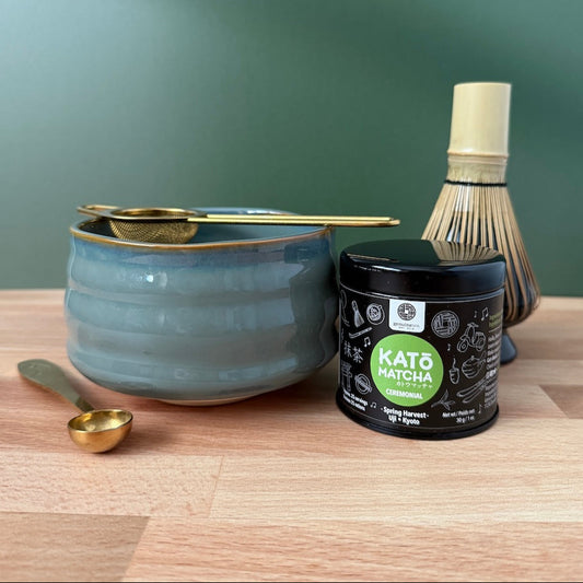 Tea-making set with a blue ceramic bowl, gold whisk, scoop, and Kato Matcha container on a wooden surface.