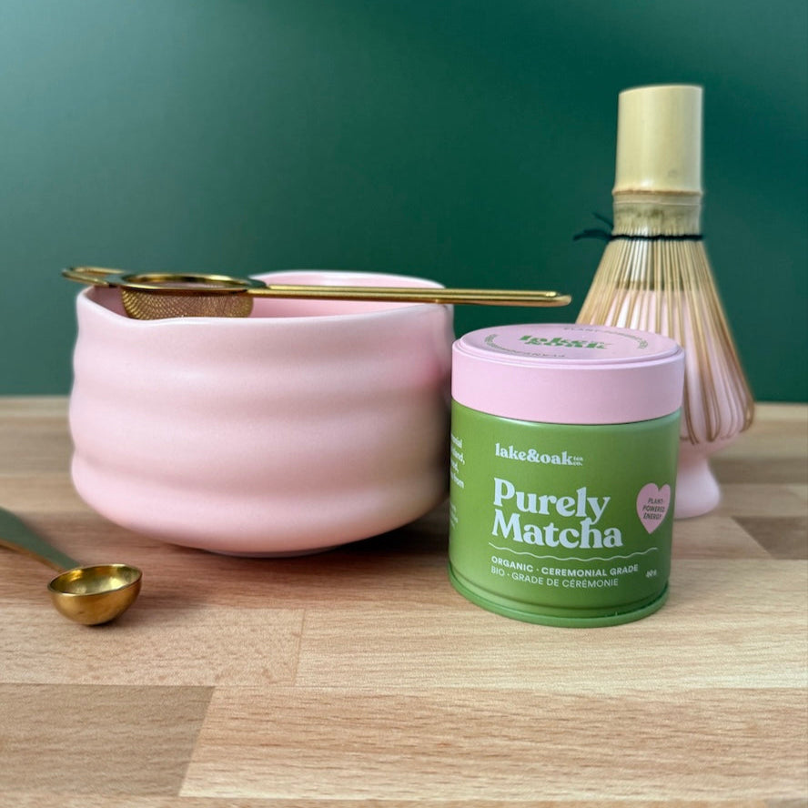Pink ceramic bowl, gold spoon, and 'Purely Matcha' container on a wooden surface with a green background.